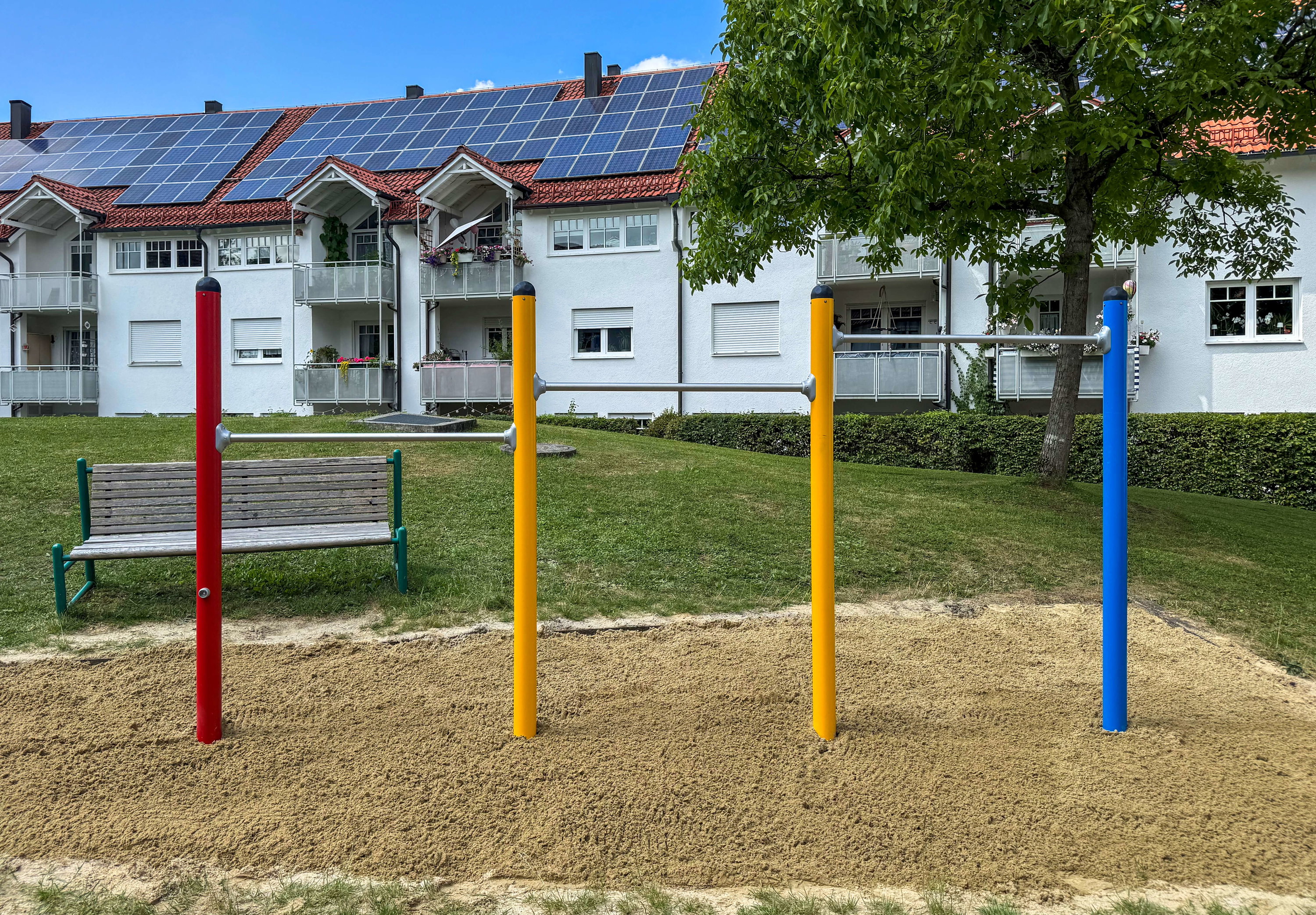 Frisch gestrichene Wohnhäuser mit Solaranlagen im Hintergrund und farbige Fitnessgeräte vorne auf Sandfläche.

alt="Farbenfrohe Fitnessstation vor weißem Mehrfamilienhaus mit Solarpaneelen"