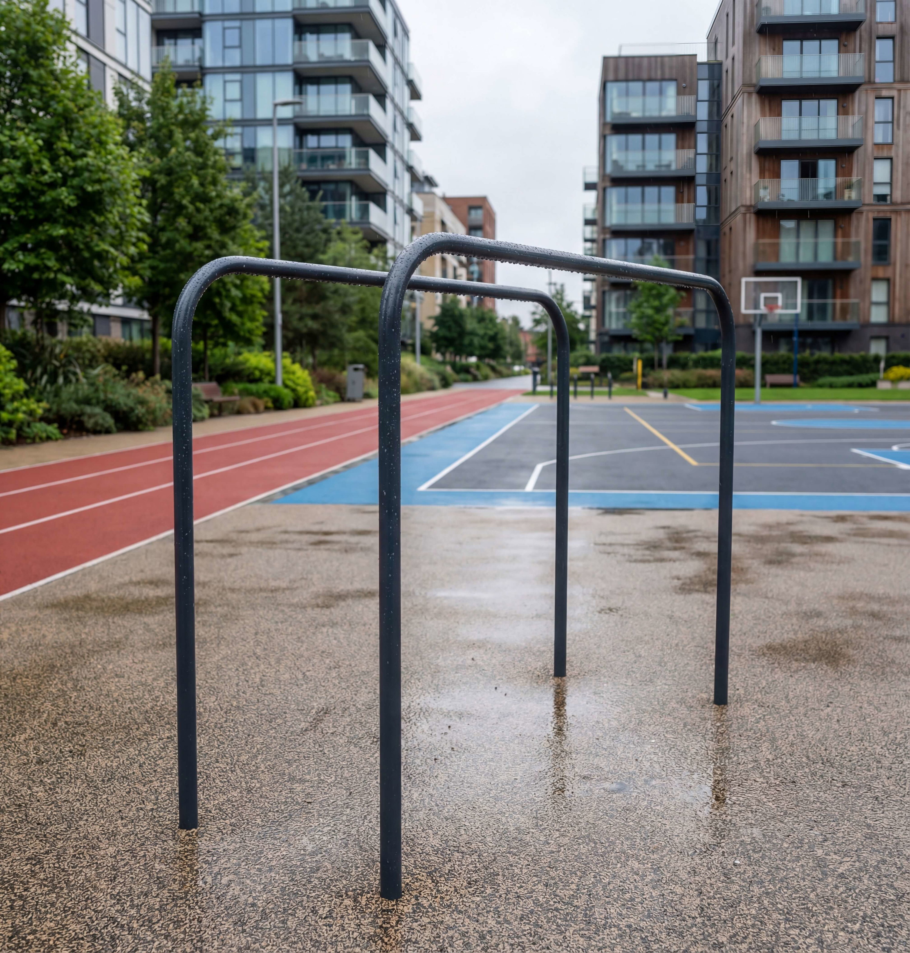 Schwarze Hantelstangen auf nasser Asphaltfläche vor Basketballplatz und Wohngebäuden.

Gymnastikgerüst im Stadtteil – feucht nach Regen, mit Laufbahn und Kletteranlagen im Hintergrund.