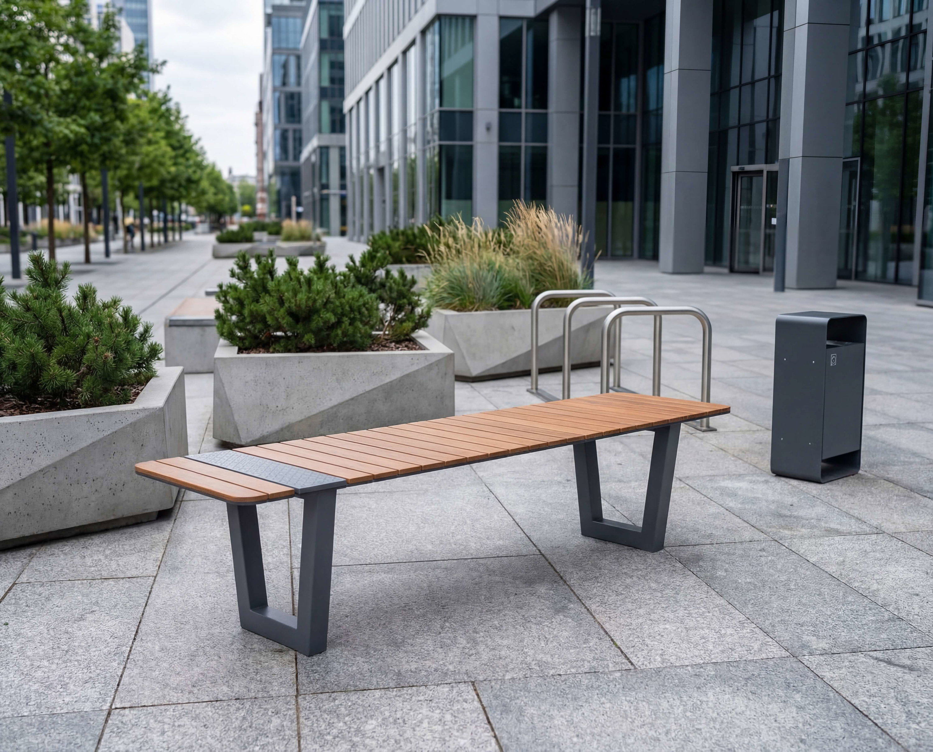 Moderne Bank mit Holzplatte auf gepflastertem Platz vor Glasfassade und Pflanzenbeeten.