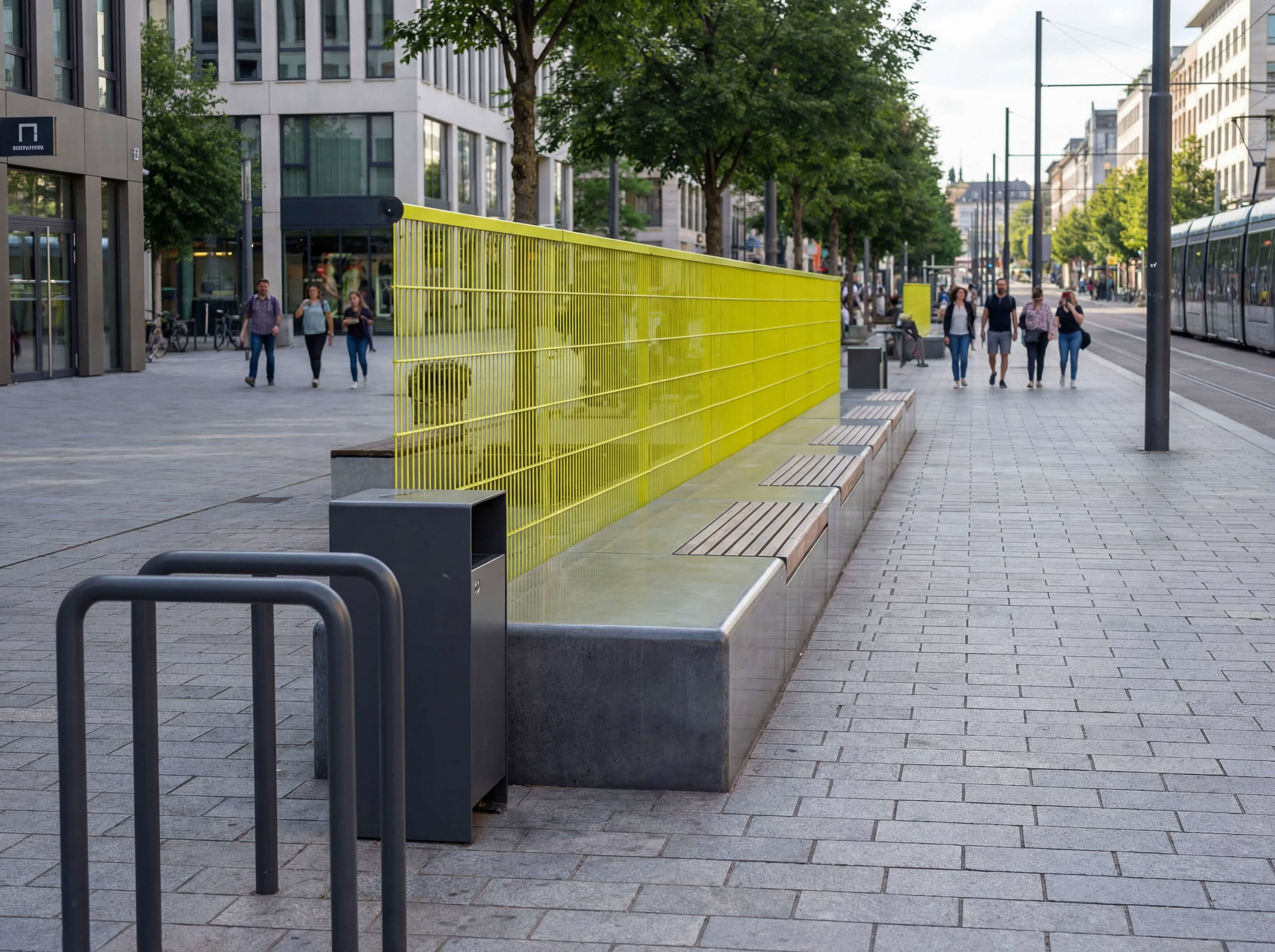 Gelber Zaun und Bank am gepflasterten Gehweg mit Passanten im Stadtzentrum.