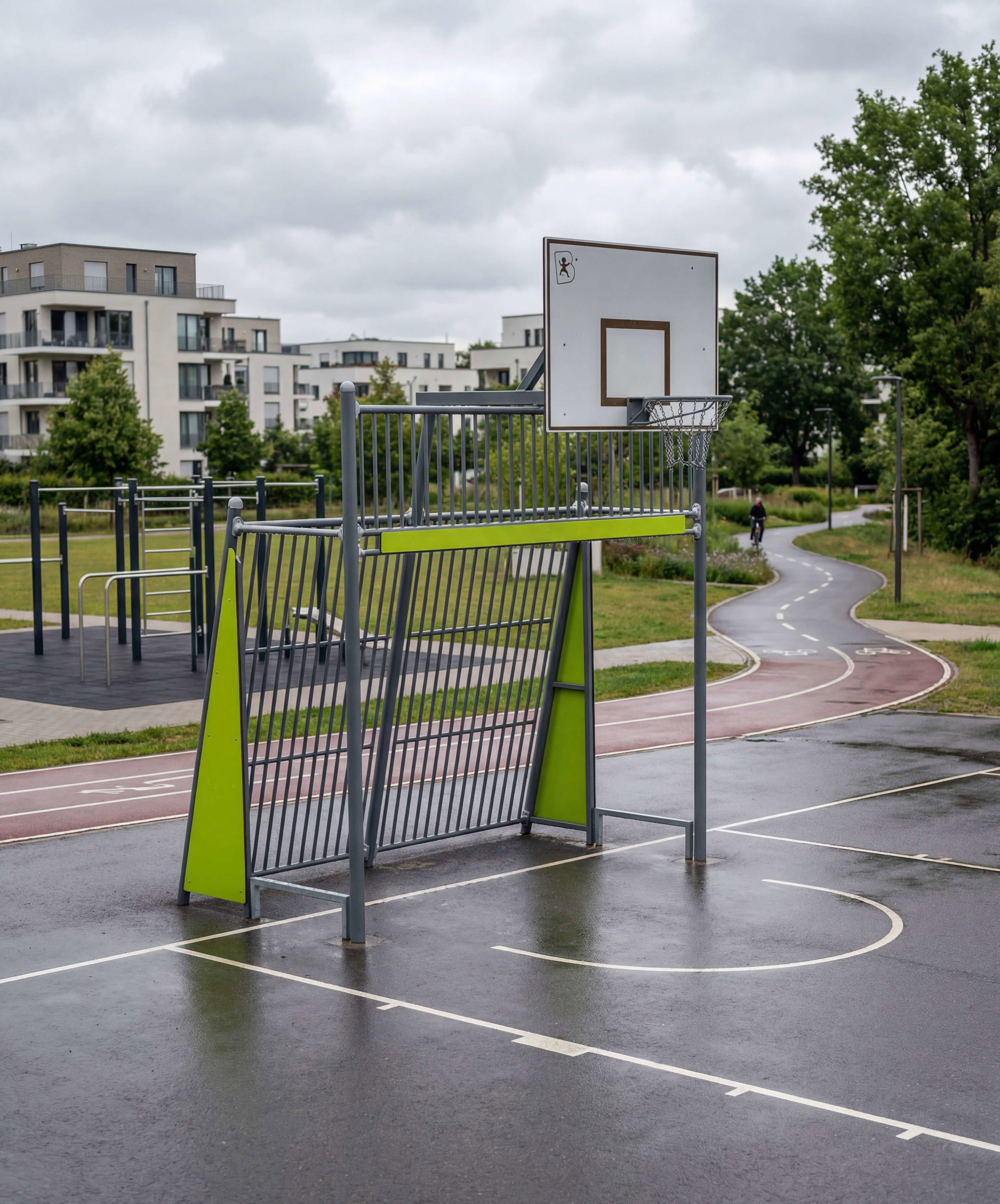 Regnerischer Basketballplatz mit grünem Korb und modernen Wohnhäusern im Hintergrund.