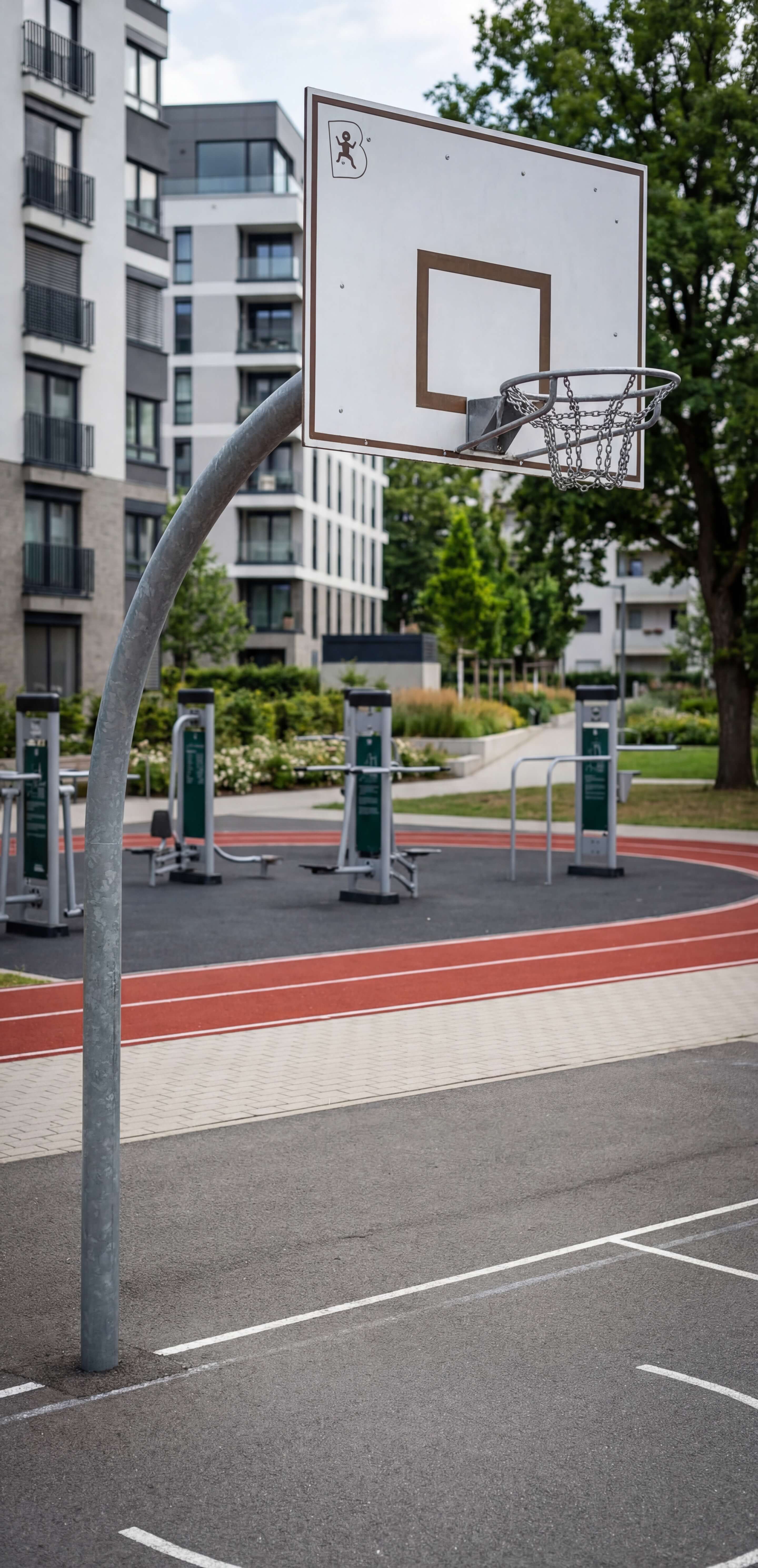 Basketballkorb auf Asphaltplatz vor Wohnhaus und Fitnessgeräten im Grünen.