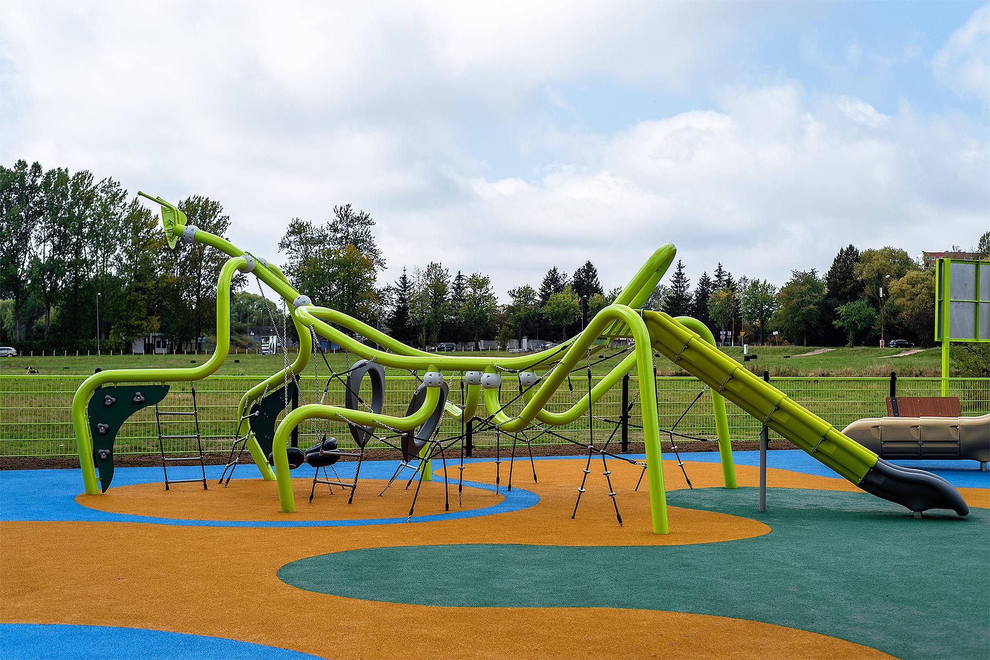 Frischer Spielplatz mit grünem Schlauch- und Rutschbereich auf buntem Boden unter blauem Himmel.