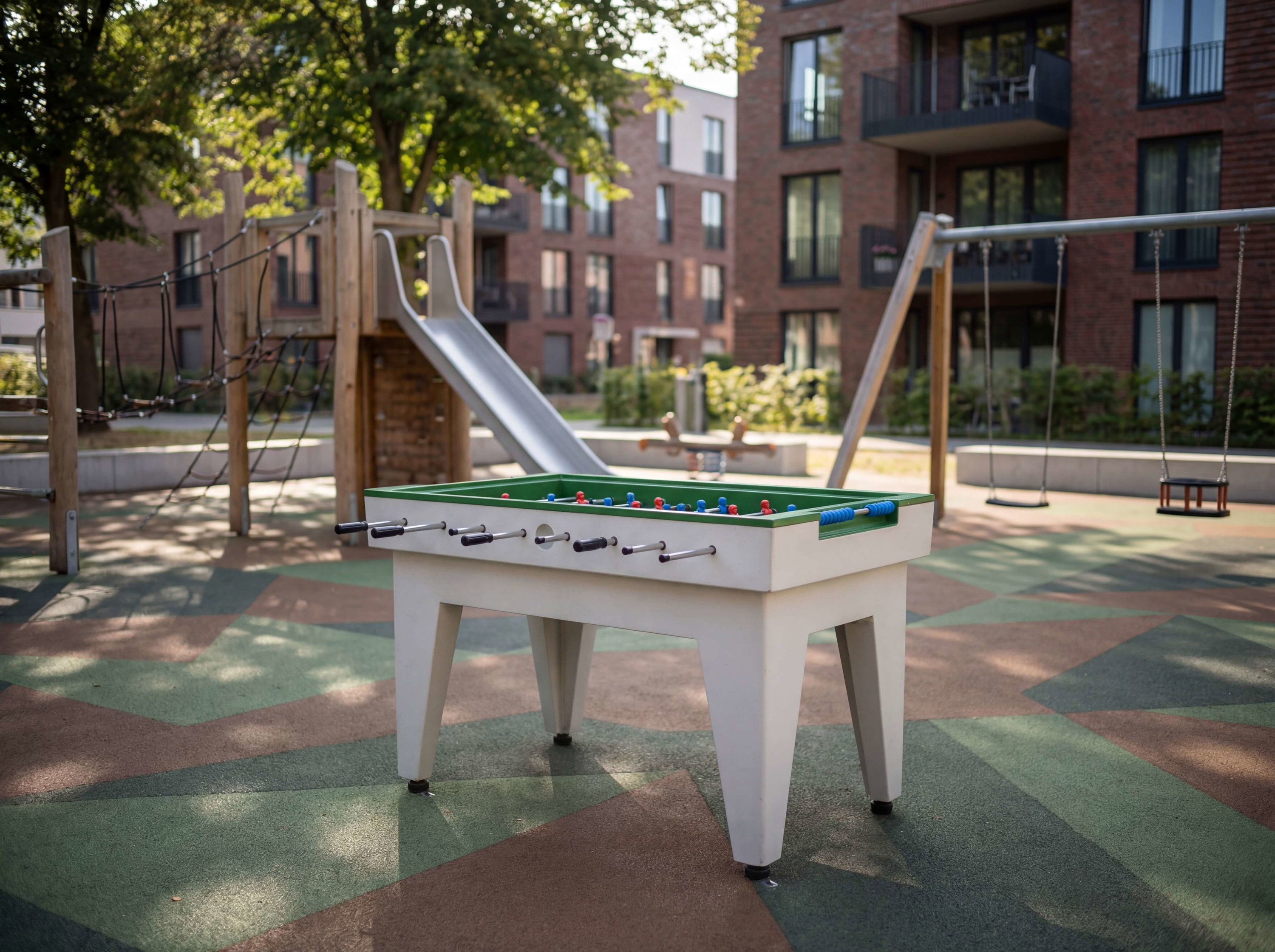 Fußballtisch im Spielplatz vor Wohnhäusern mit Rutschbahn und Schaukeln unter Bäumen.