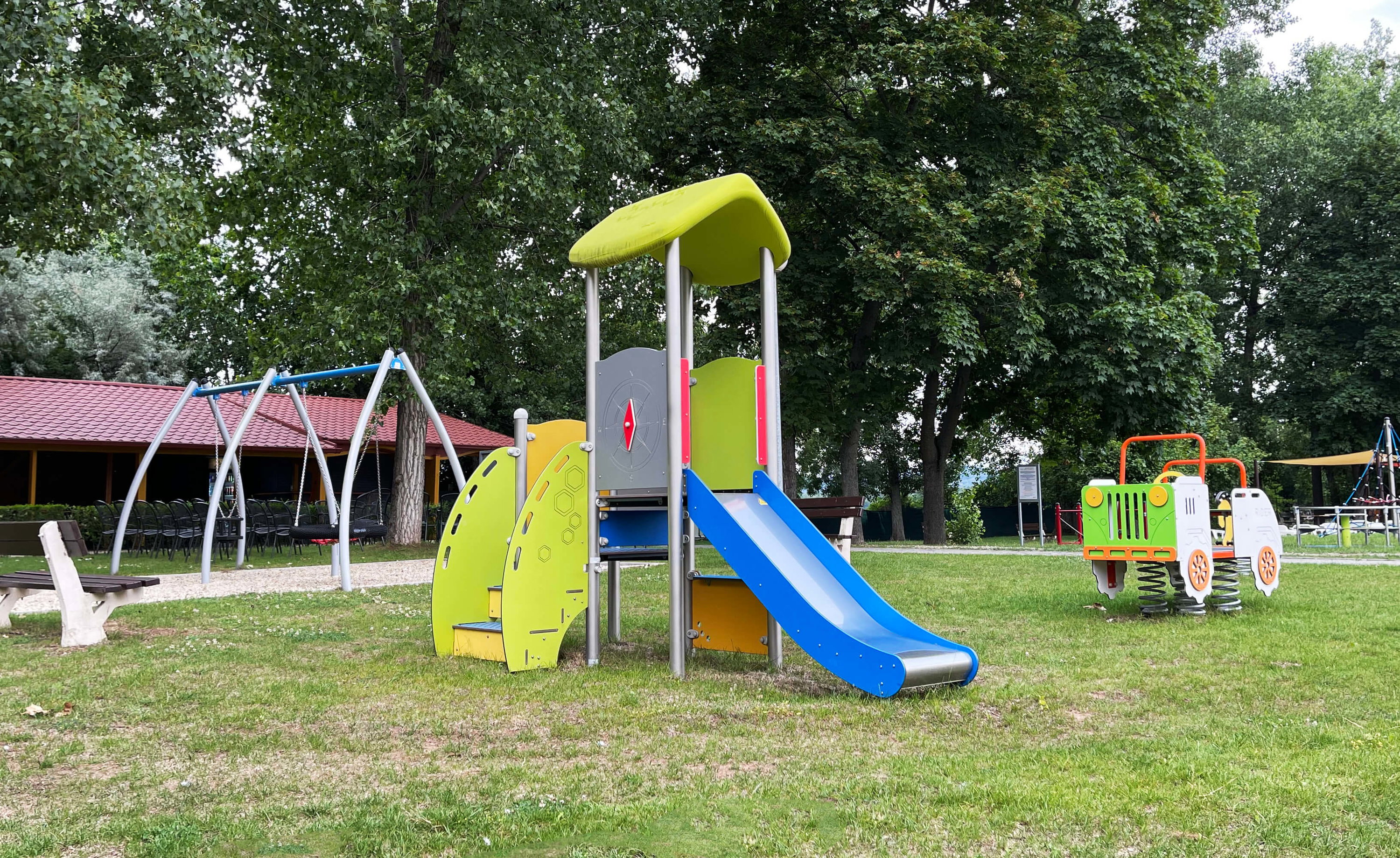 Bunte Spielplatzanlage mit Rutsche und Schaukel im grünen Park unter Bäumen.