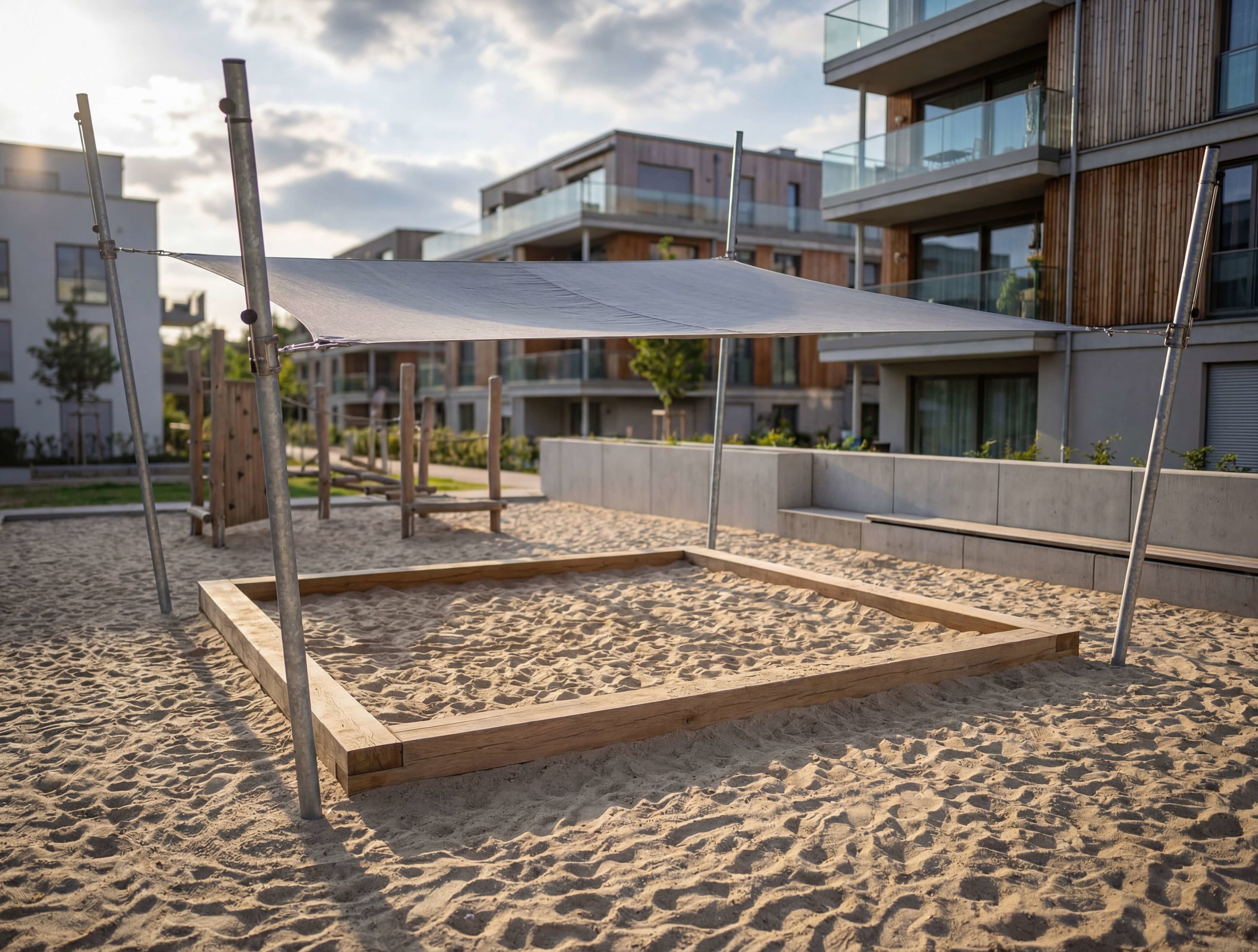 Spielplatz mit Sandkasten und Sonnensegel vor modernem Wohnhaus im Abendlicht.
