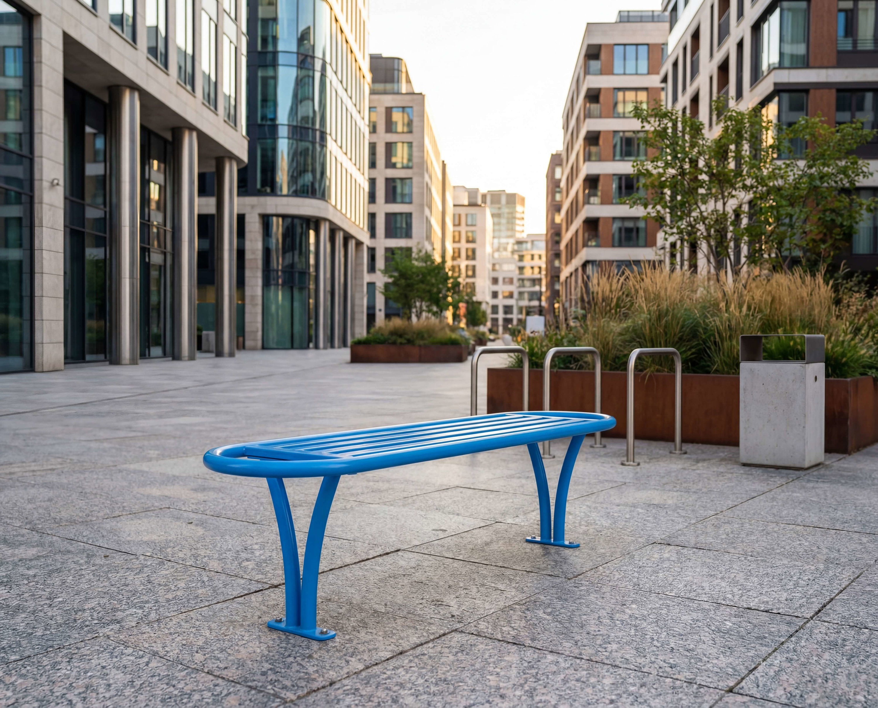 Blauer Stahlbänkchen auf gepflastertem Platz zwischen modernen Hochhäusern und Pflanzenbeeten.