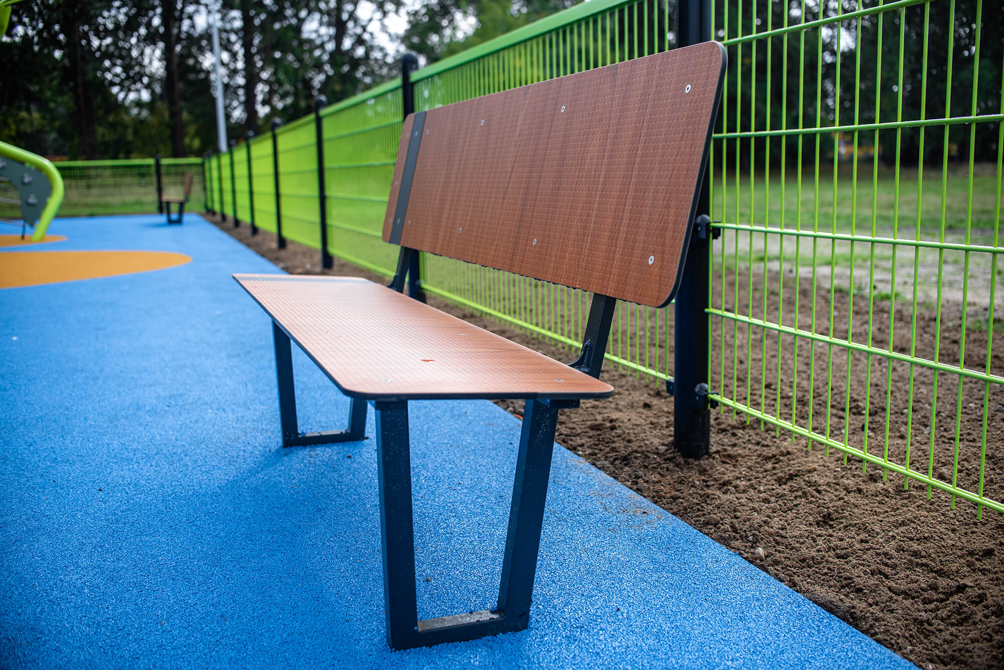 Holzbank auf blauem Spielplatzboden neben grünem Zaun im Park.