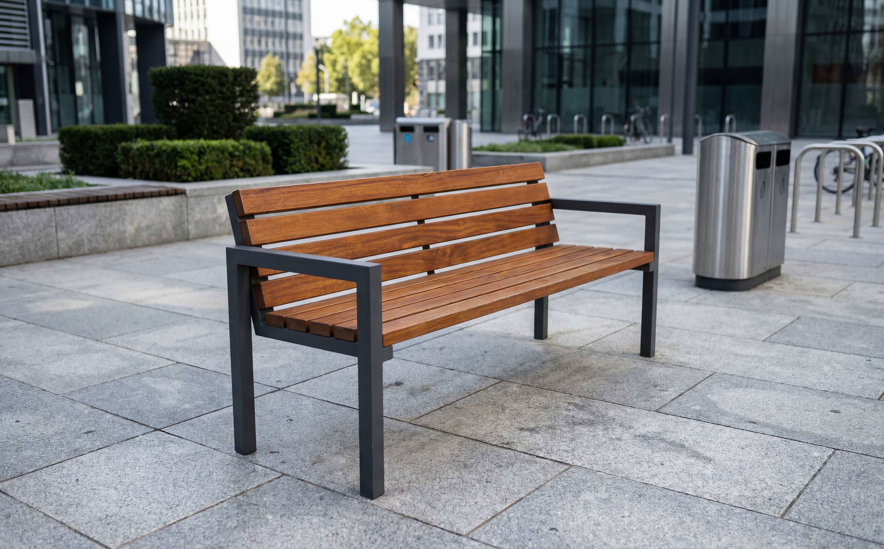 Holzbank mit Metallgestell auf gepflastertem Platz vor modernen Gebäuden und Bäumen.