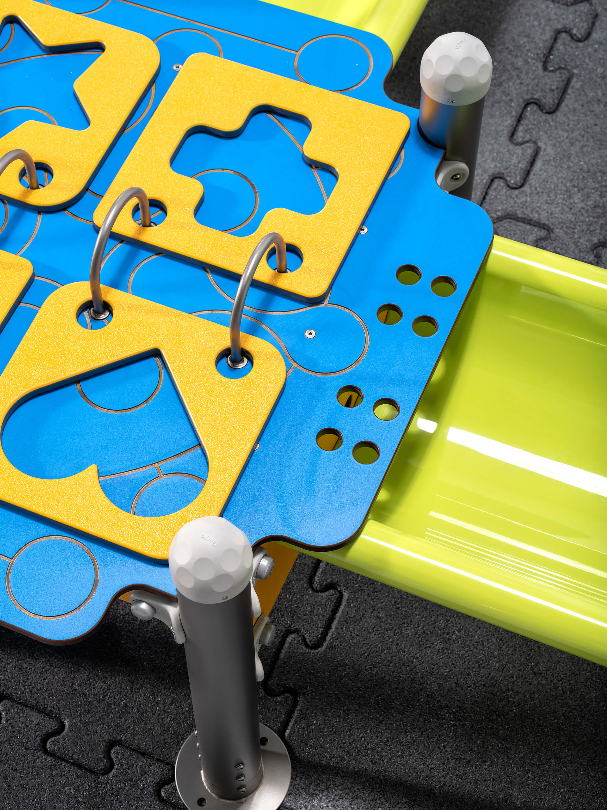 Bunte Spielplatte mit gelben Formen und Metallstangen auf schwarzem Gitterboden.