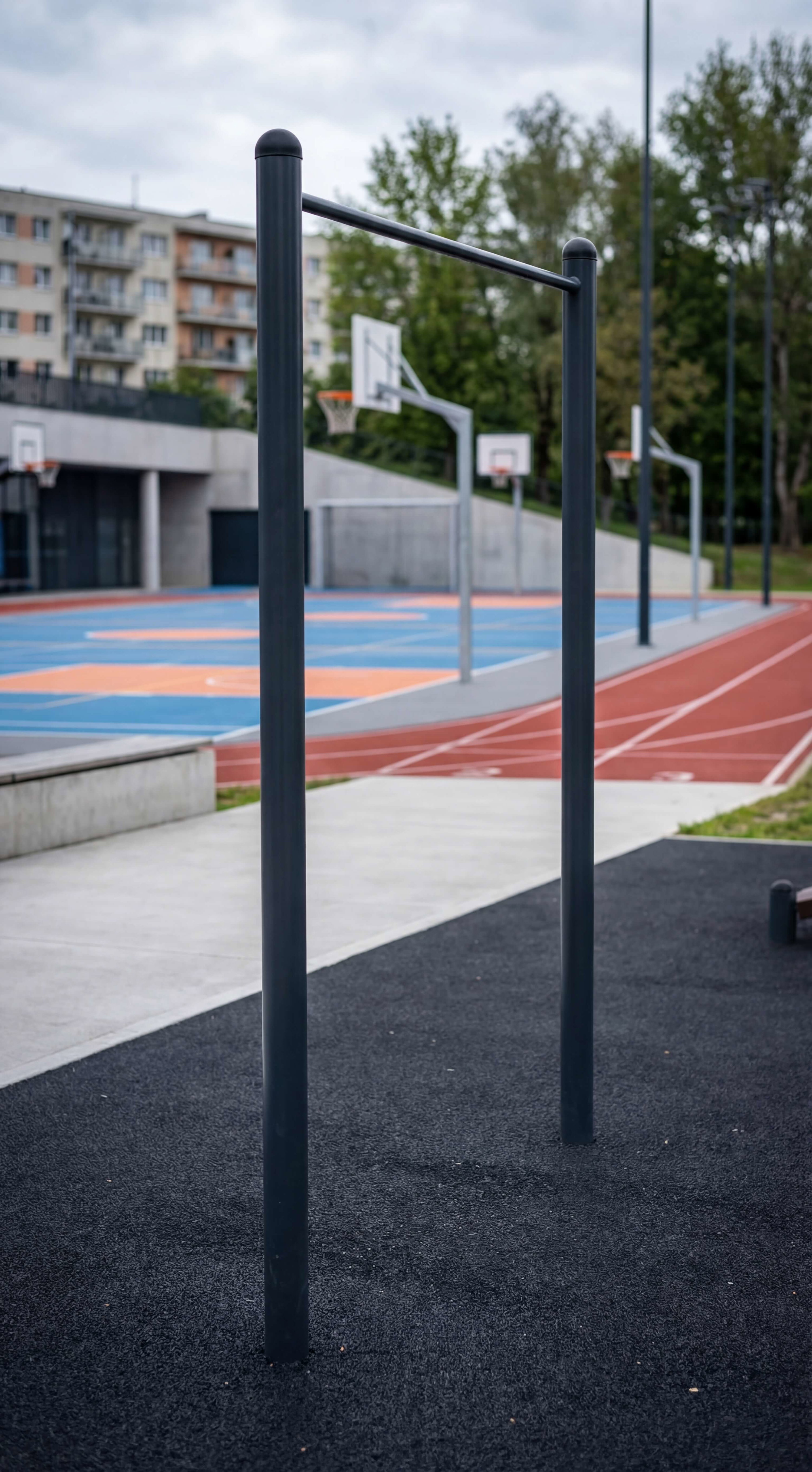 Schwarze Kletterstange auf Asphaltboden vor Basketballplatz und Wohnhaus im Hintergrund.