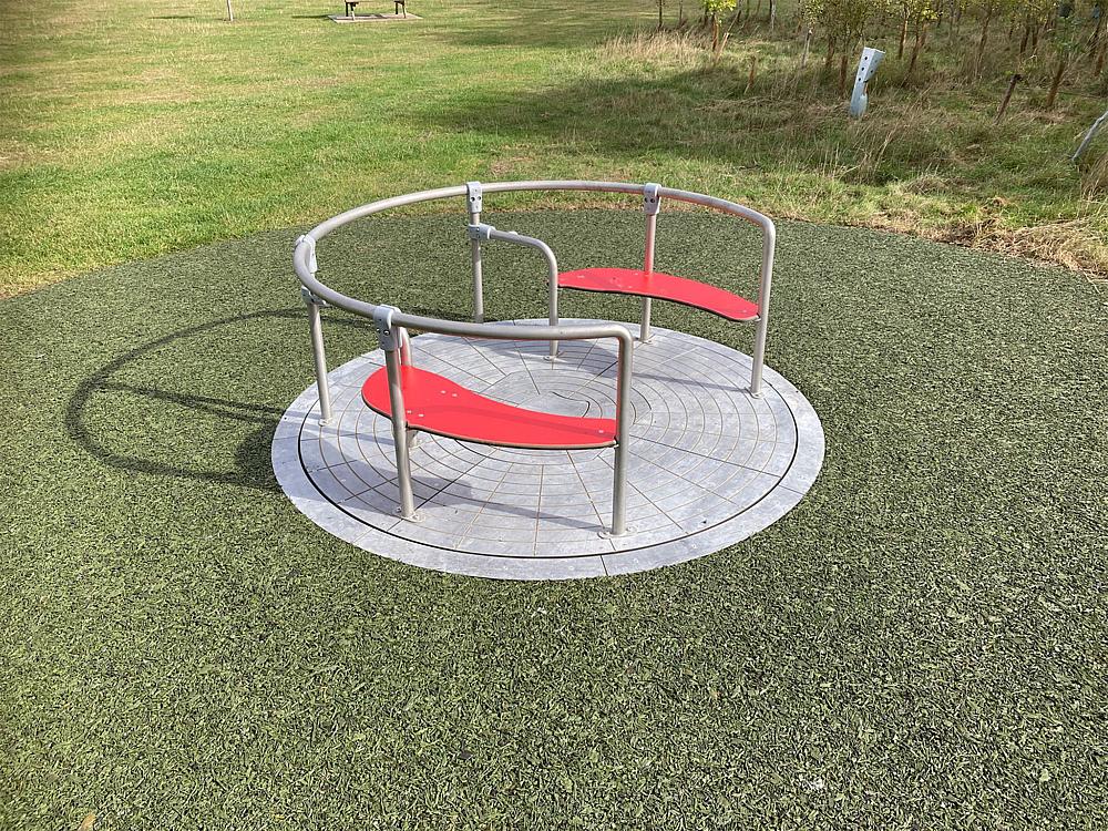 Roter Drehplatz mit Metallgeländer auf grünem Kunstrasen im Park.