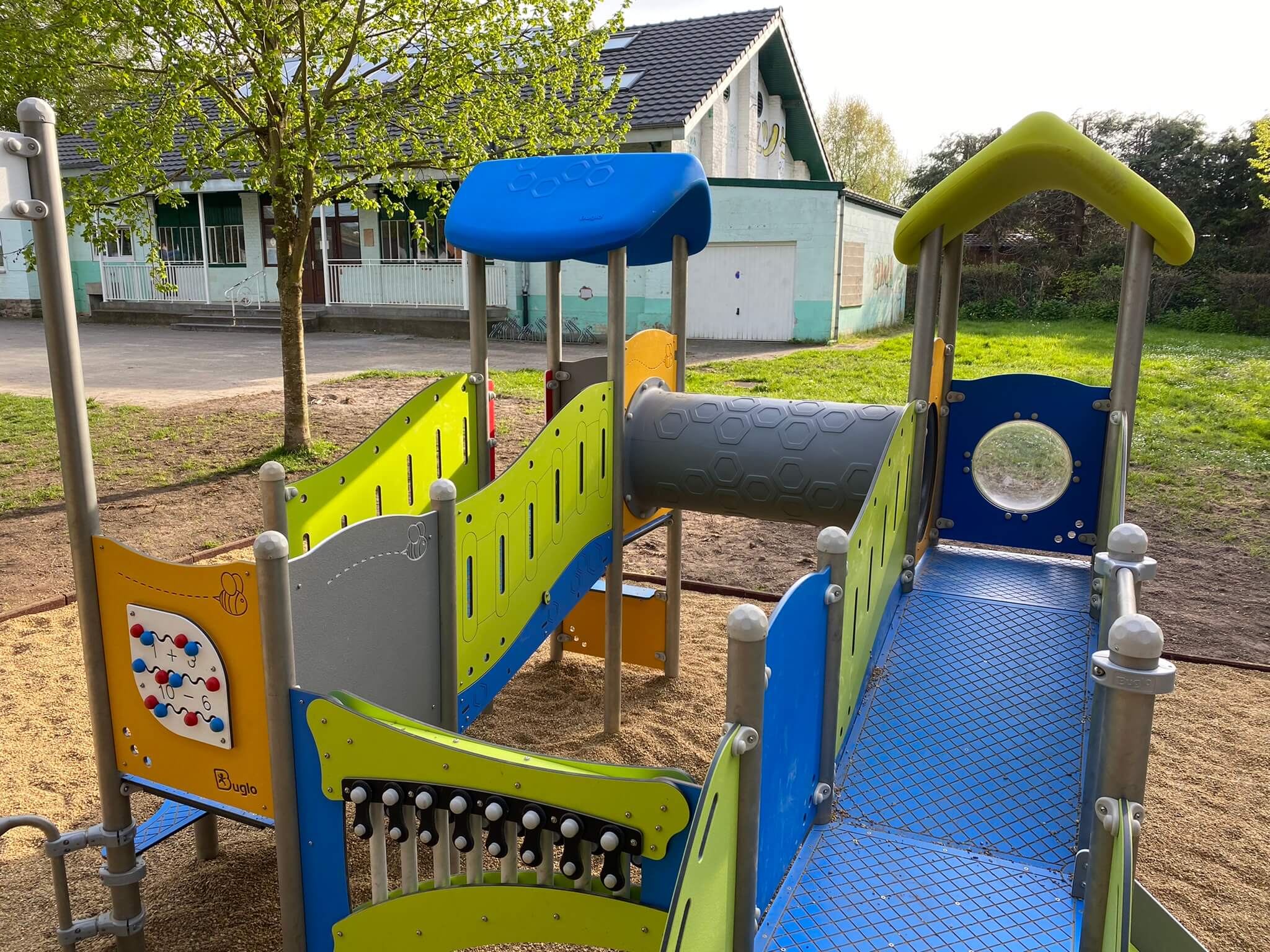 Bunte Spielplatzanlage mit Rutsche und Kletterelementen vor einem Schulgebäude im Freien.