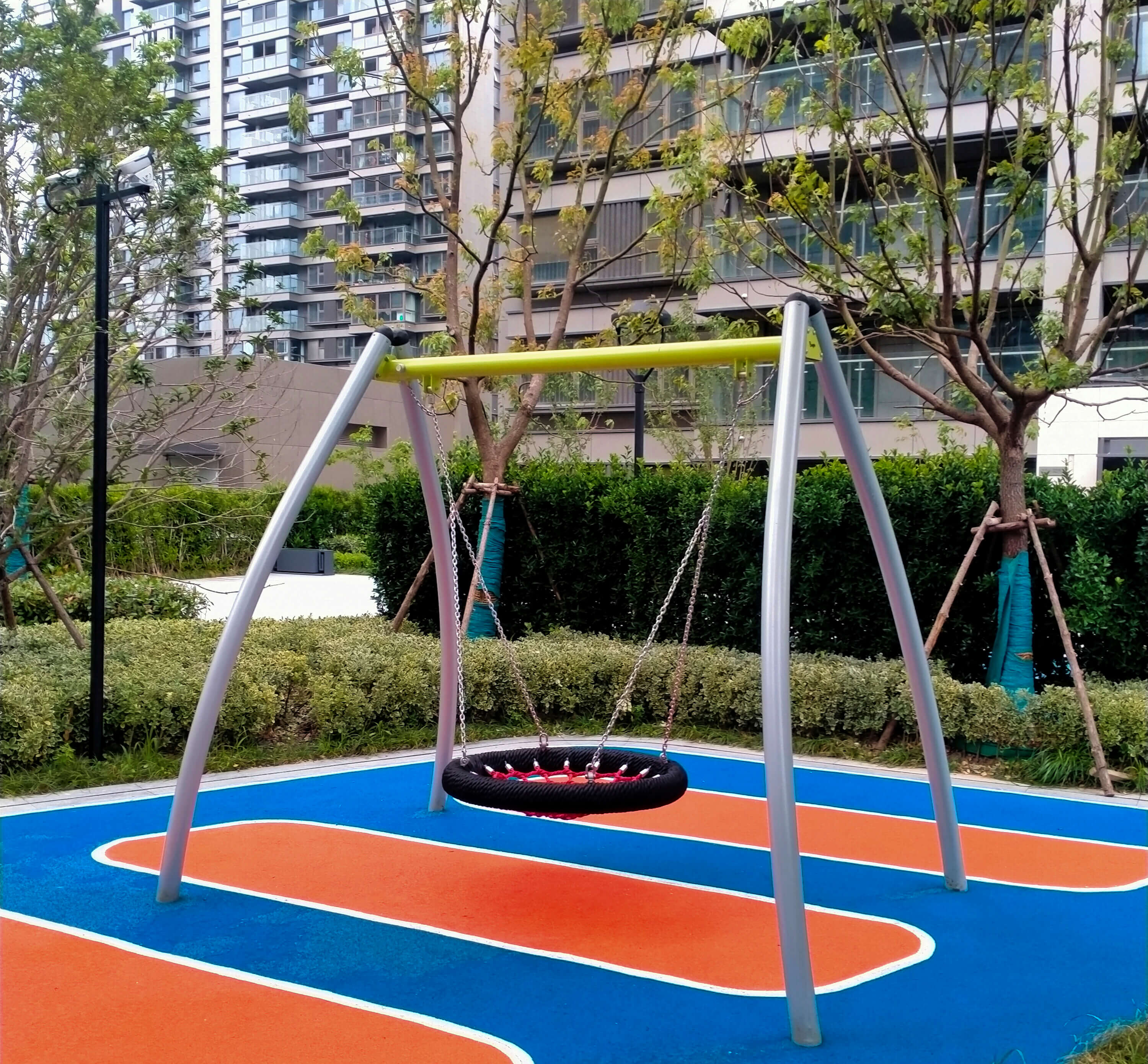 Schaukeln im modernen Spielplatz vor Hochhäusern mit buntem Boden und Bäumen.