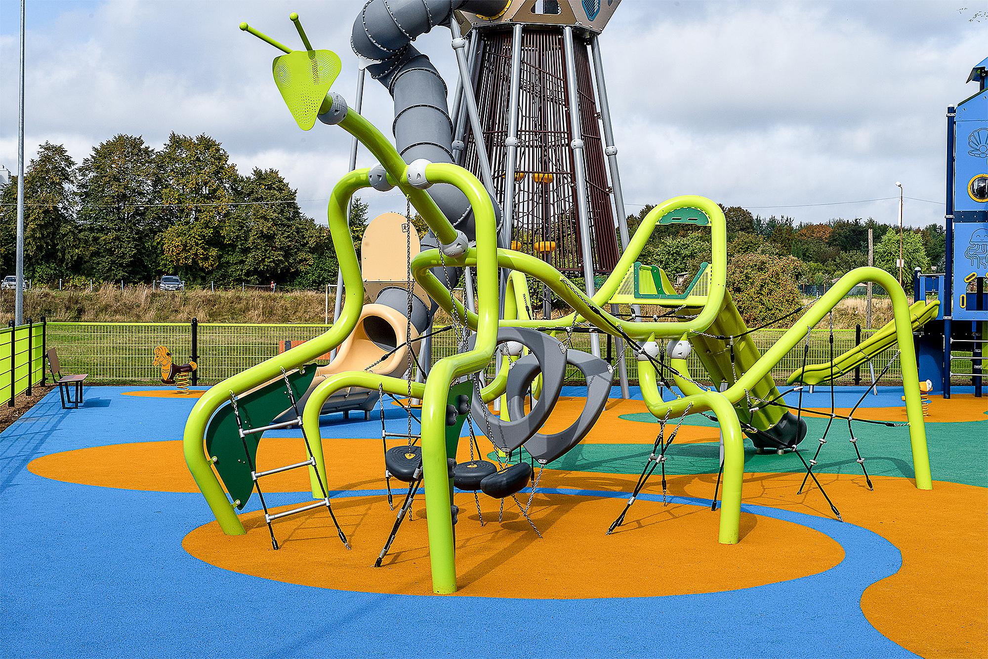 Bunte Spielplatzanlage mit Rutschen und Klettergerüsten auf blau-orange gepolstertem Boden unter bewölktem Himmel.