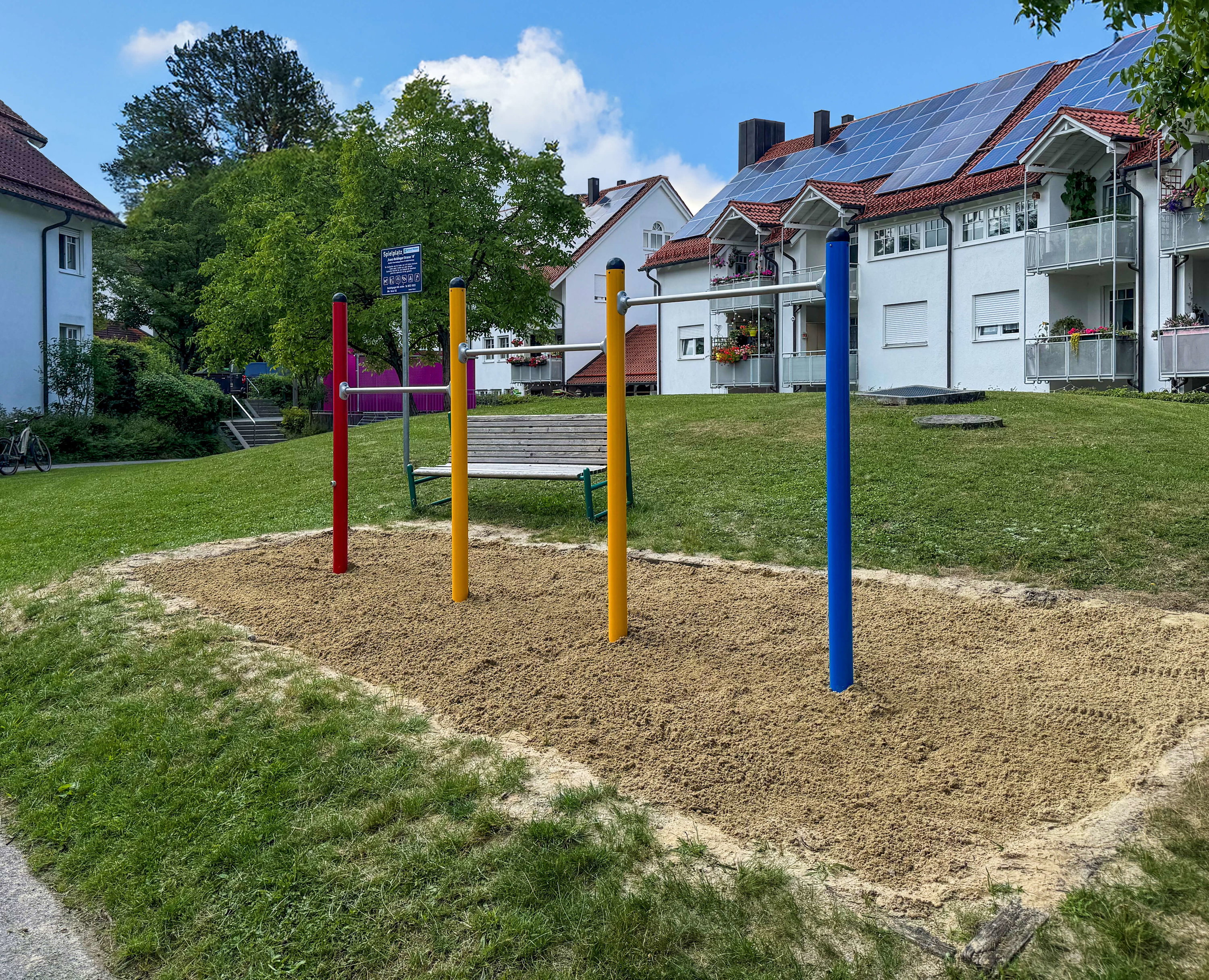 Farbiger Fitnessbereich im Grünen vor Wohnhäusern mit Solaranlagen unter blauem Himmel.
