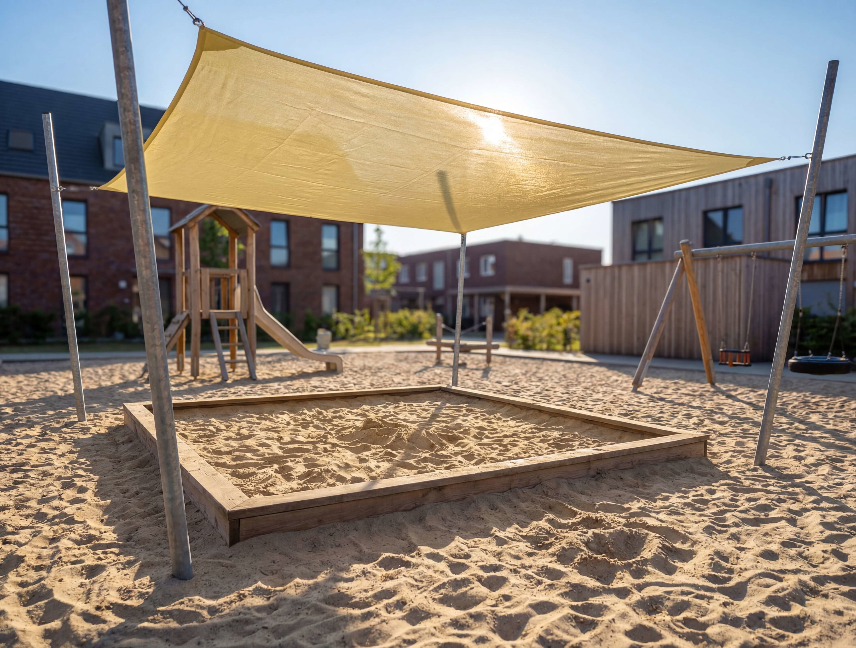 Spielplatz mit Sandkiste und Sonnensegel vor Wohnhäusern im warmen Licht der untergehenden Sonne.