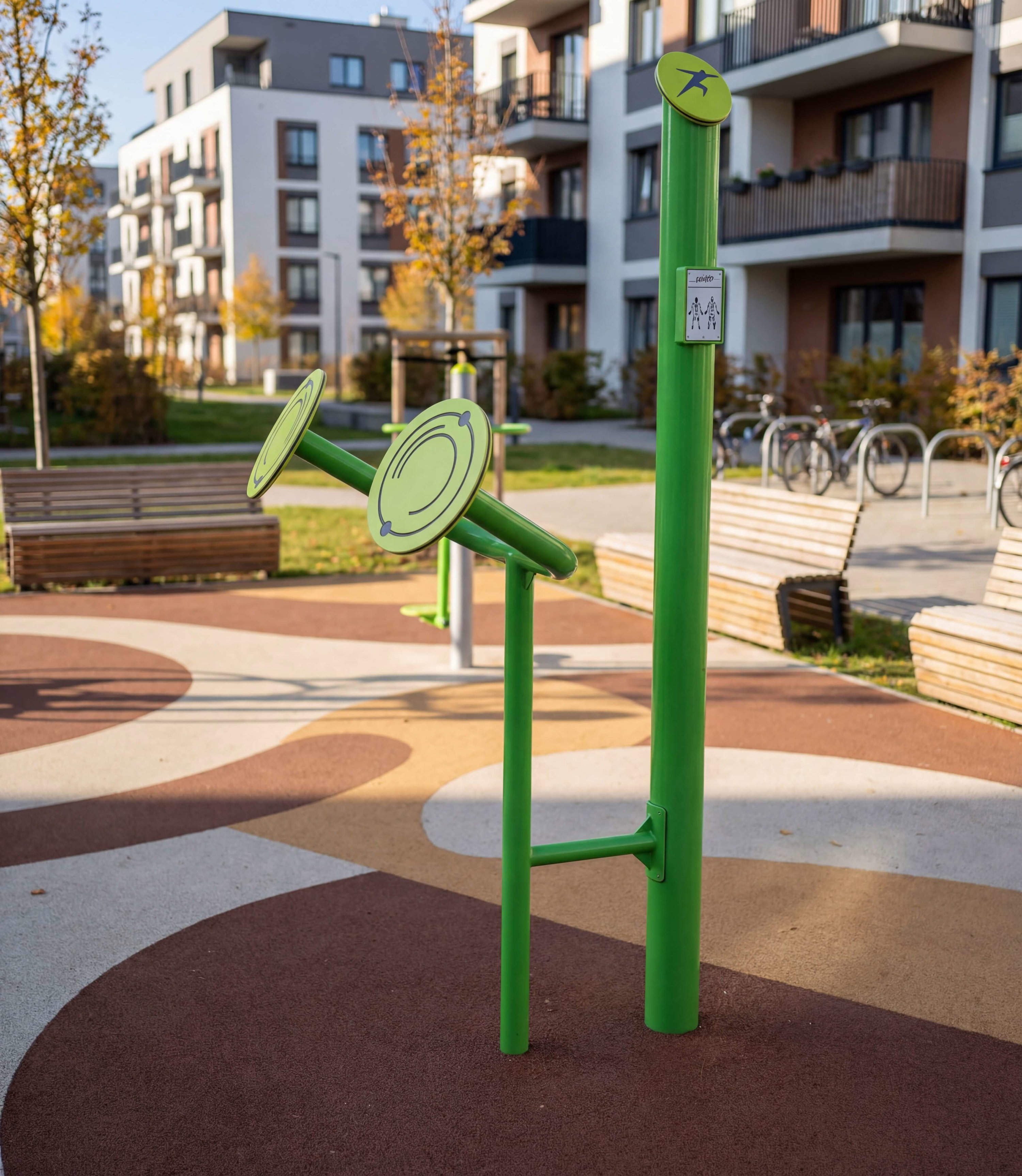 Grünes Fitnessgerät im städtischen Park vor Wohnhäusern und Fahrradständern.