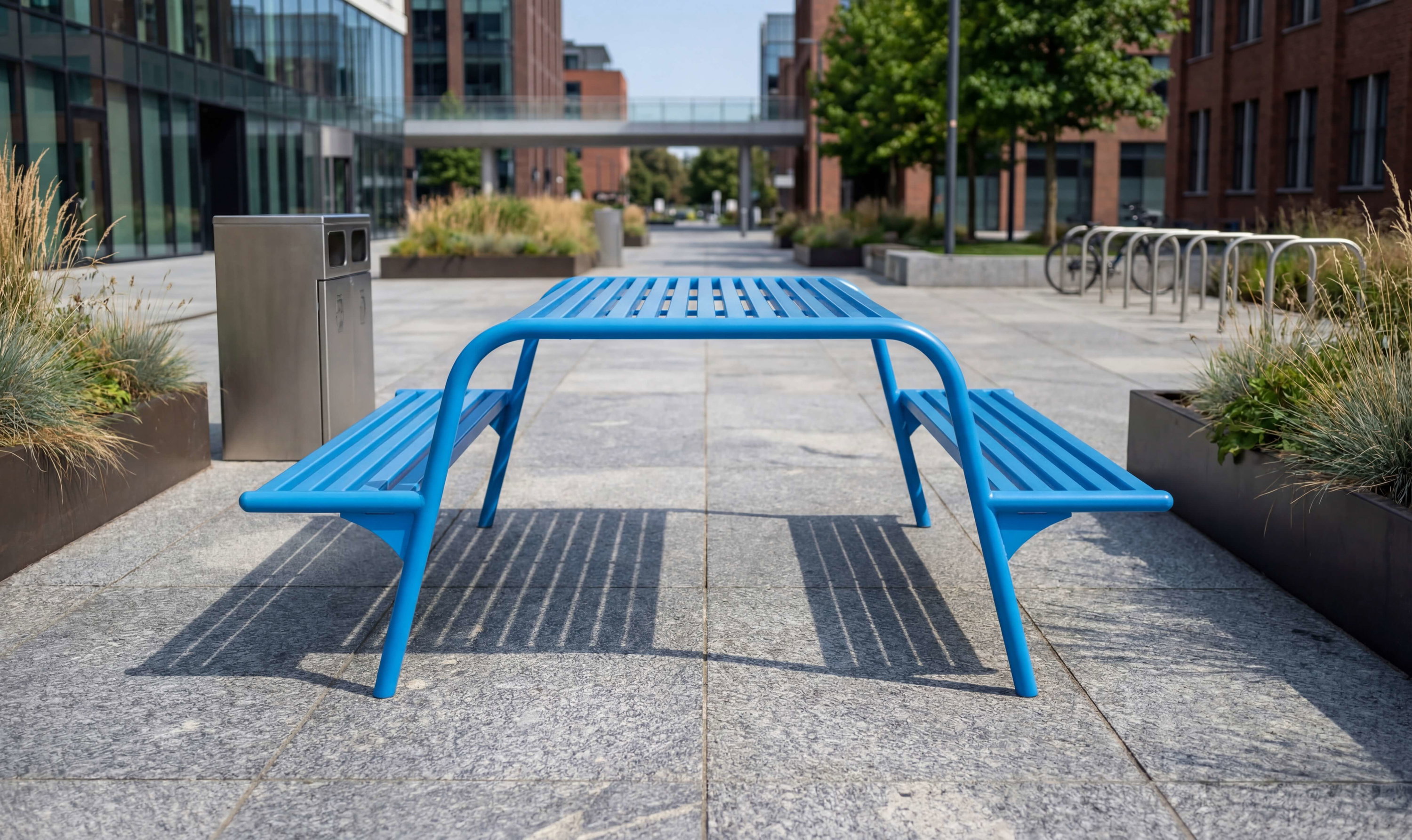 Blauer Metalltisch mit Bänken auf Steinplatten vor modernen Gebäuden und Grünanlagen.