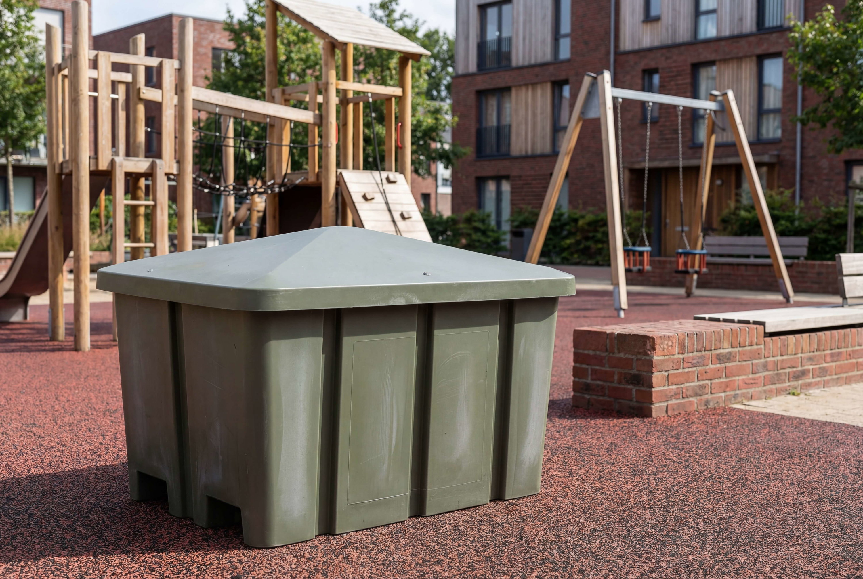 Grüner Spielplatzkasten auf rotem Kies vor Holzspielgeräten und Wohnhäusern.