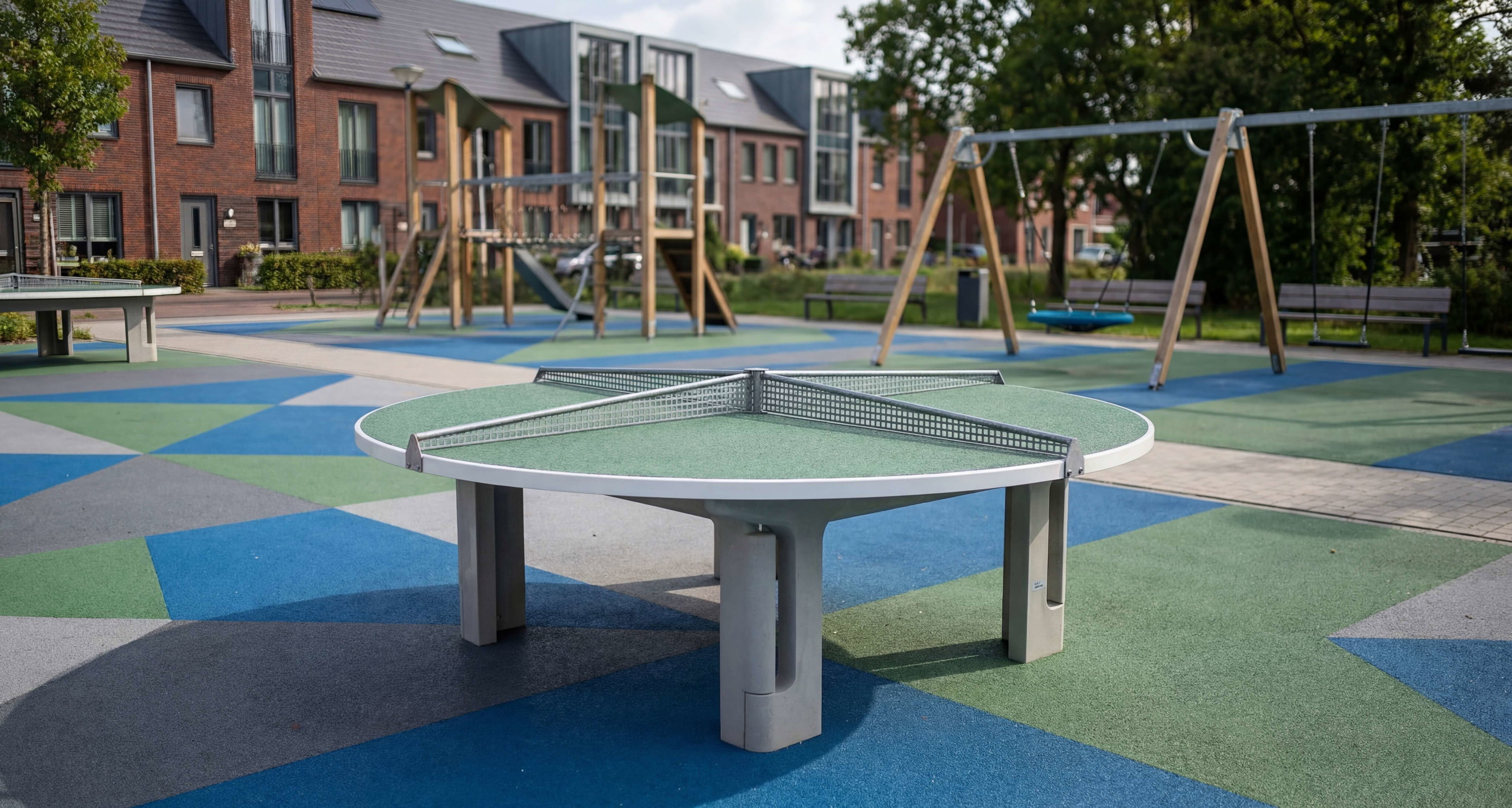 Runder Tischtennis-Tisch auf buntem Spielplatz vor Wohnhäusern und Schaukeln im Freien.