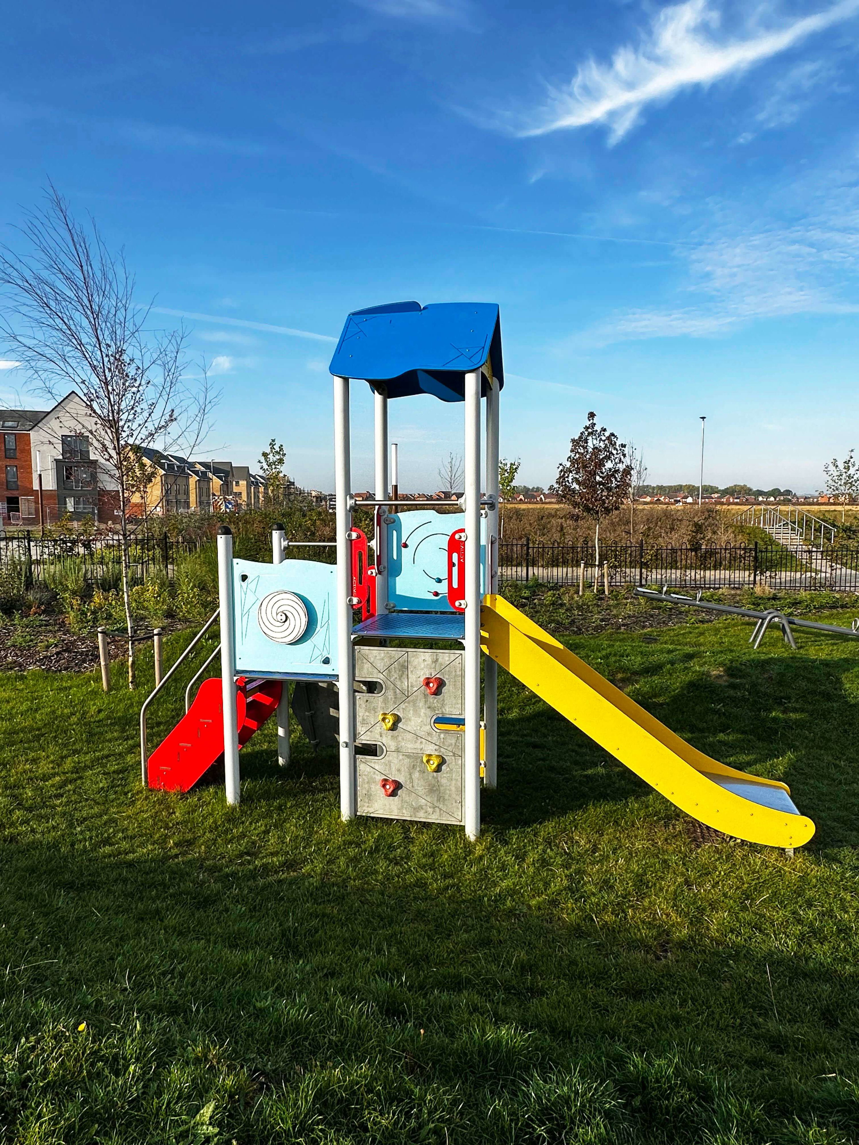 Bunte Spielplatzanlage mit Rutsche und Kletterwand auf grünem Rasen unter blauem Himmel.